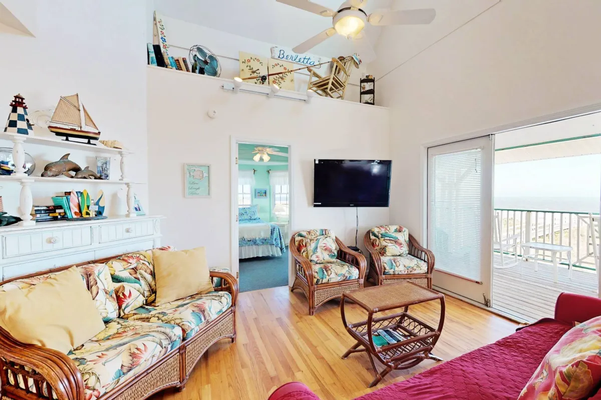 A casual and inviting living room featuring comfortable floral-patterned sofas, decorative shelves with nautical accents, and expansive windows that open onto a balcony with ocean views. The space is clearly designed for socializing or relaxing, with ample natural light streaming in.