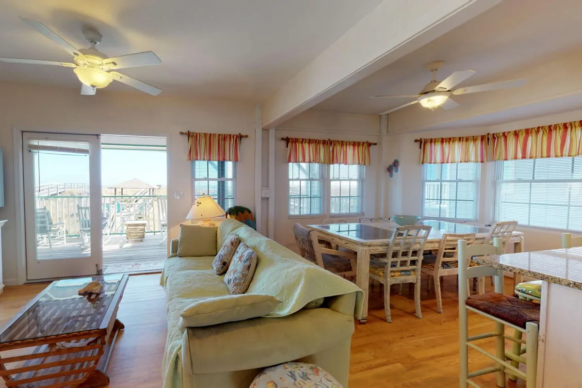 An open-plan living and dining area with a comfortable sofa, large windows offering outdoor views, and a dining table perfect for group activities. The room is bathed in sunlight and designed for both relaxation and lively gatherings.
