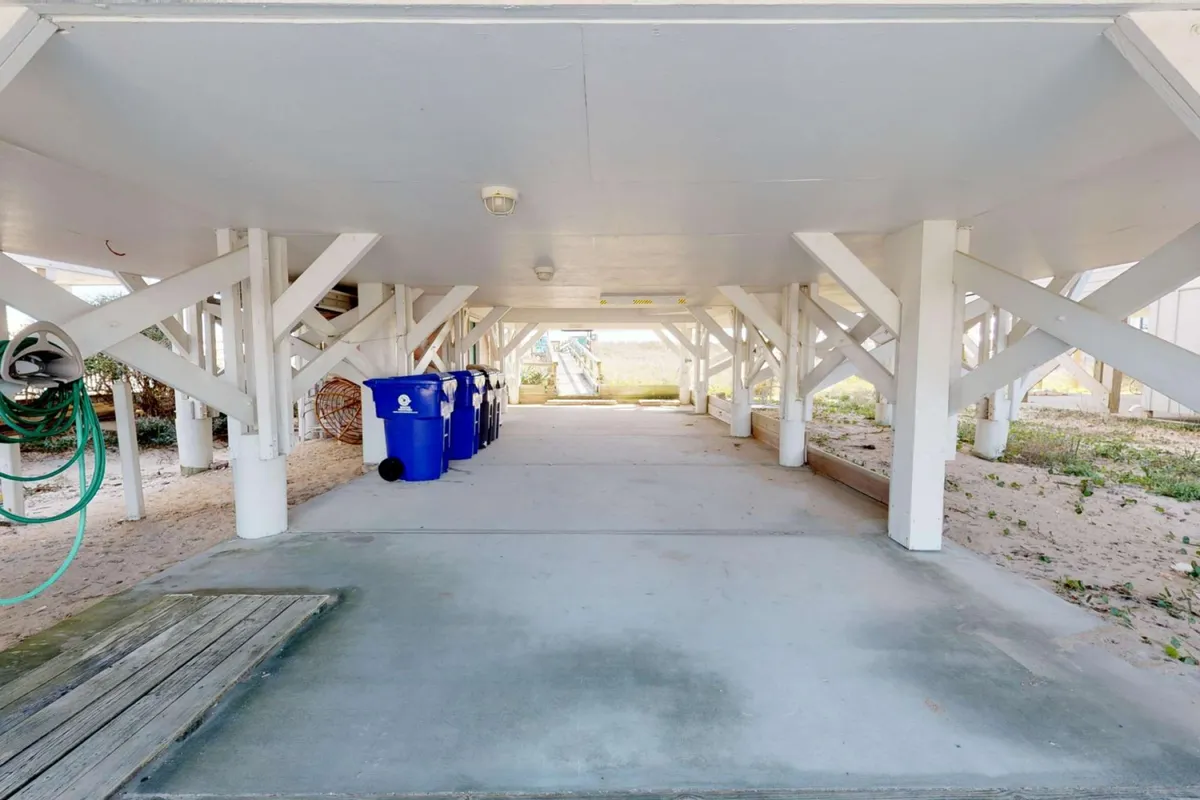 The shot shows an exterior sand-level area under the house, with visible support beams, blue trash bins, and an open sand area, providing ample space for storing beach gear and equipment.