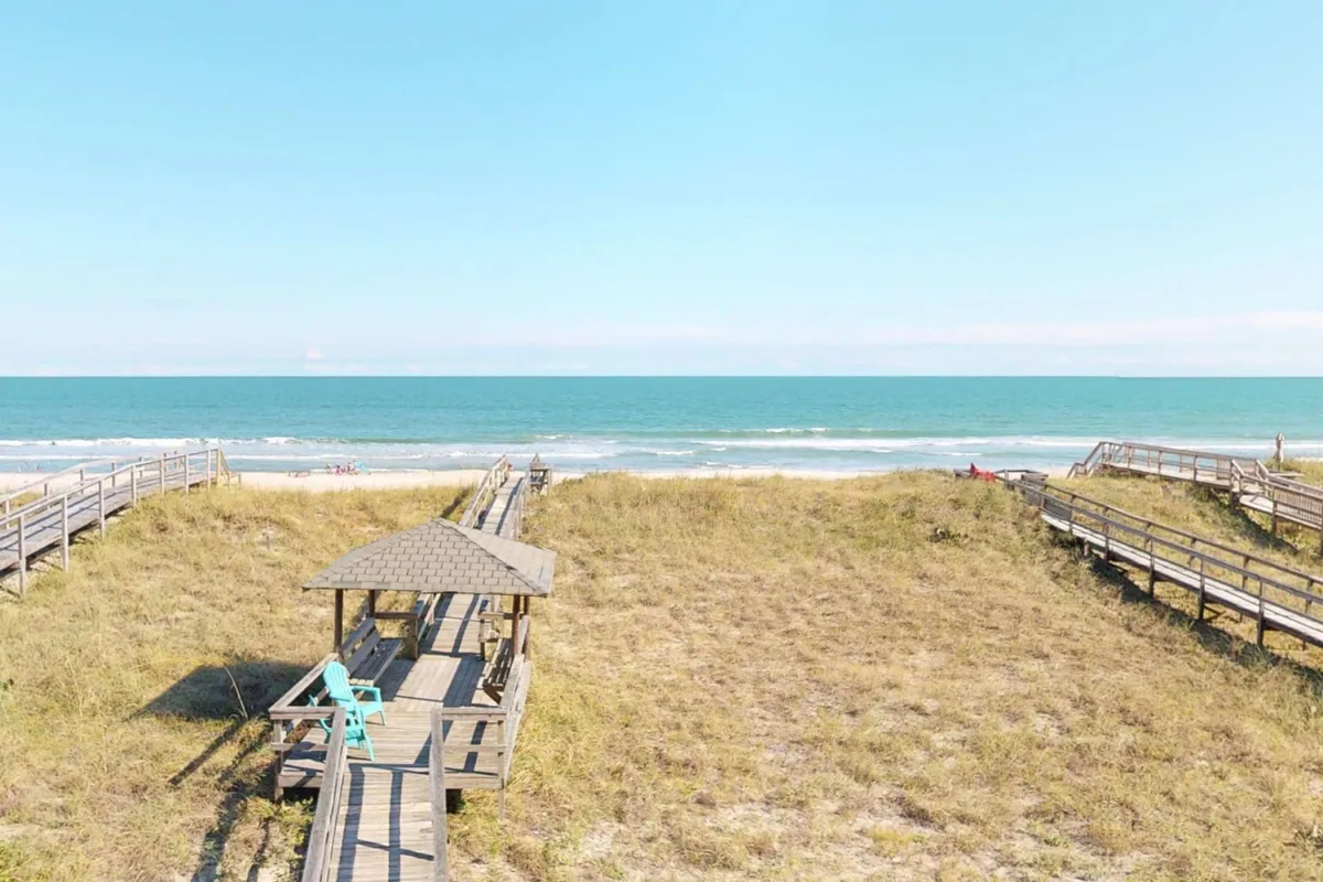 A wooden boardwalk leads through dune grass to a charming beach gazebo, with the ocean waves in the distance. The relaxed atmosphere suggests activities like beachcombing, picnicking, or simply watching the sunset.
