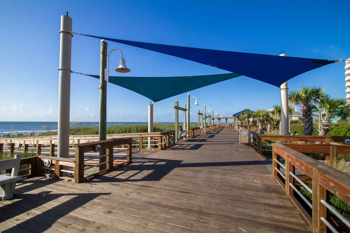 A lively wooden boardwalk stretches along the shore, shaded by blue and green sails with views of the beach and palm trees. Lampposts line the path, and the ocean is visible beyond, inviting visitors to walk, bike, or simply relax by the seaside.