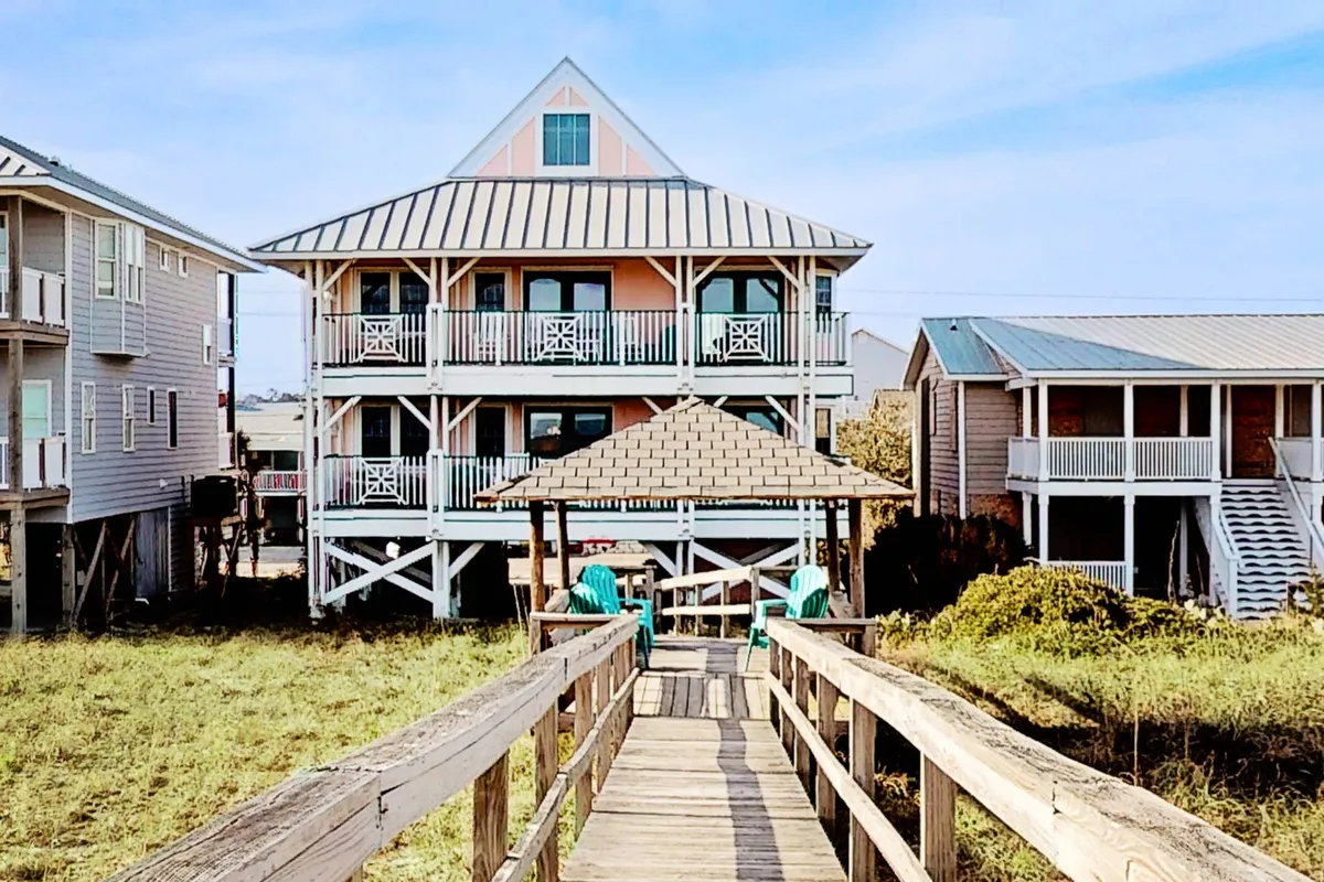The hero image showcases the grand exterior of LegaSea Beach House, with its inviting, spacious porches overlooking the ocean. A charming gazebo sits along a private pathway leading from the house, perfect for gatherings or morning coffee while watching the sunrise. The scene is bathed in warm, cheerful light, creating a welcoming and relaxing atmosphere.