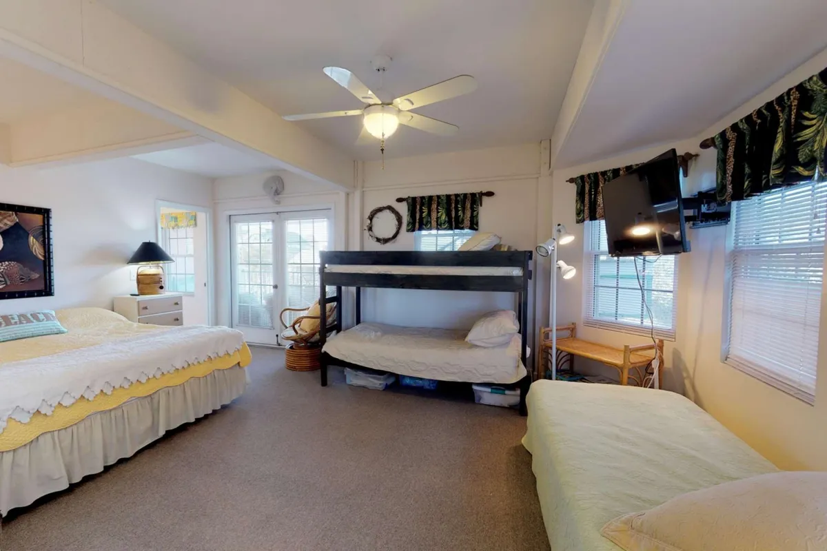 A family-friendly bedroom setup with a large bed, a bunk bed for children, windows letting in morning sunlight, a ceiling fan, and a wall-mounted television. The space is arranged to accommodate both adults and kids, with inviting bedding and a cozy atmosphere.