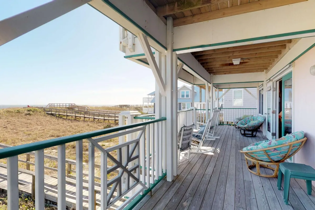 A wooden deck with rocking and woven chairs overlooks the beach, with a pathway leading toward the ocean visible beyond the railing. The scene invites relaxation and group gatherings while taking in sunset or ocean views.