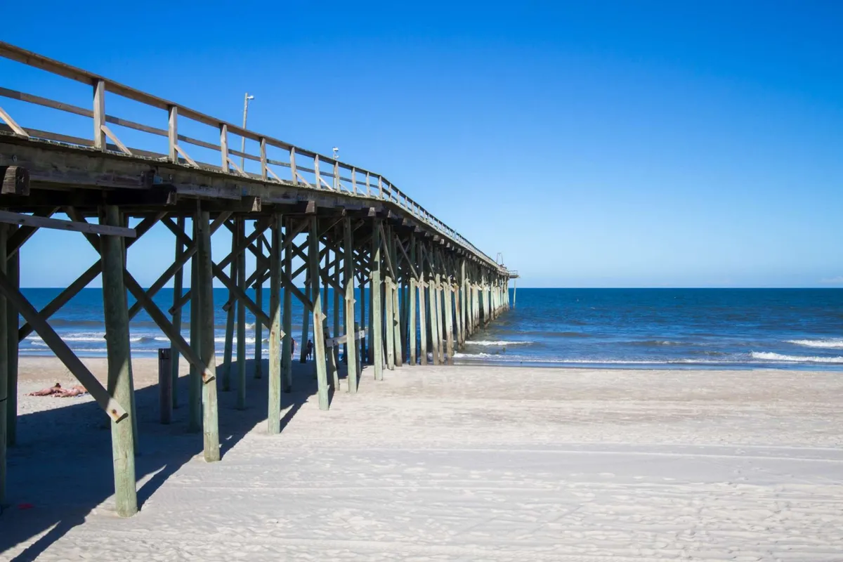 A long wooden pier extends over the ocean waves with a sandy beach below. The peaceful setting is perfect for fishing, enjoying a walk over the water, or watching sunrises and sunsets.