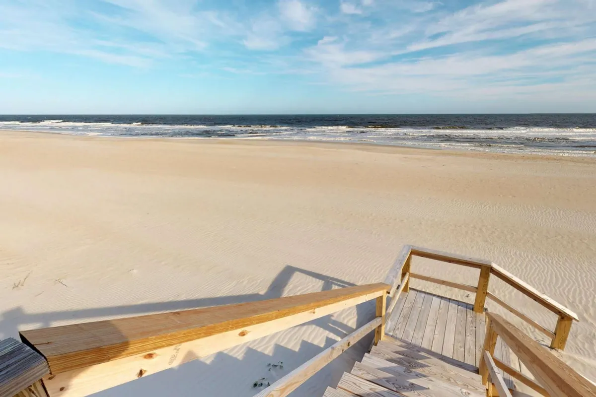 Expansive sandy beach with wooden stairs leading down, ocean waves in the background, and a vast blue sky. The image evokes a sense of adventure and play for families and groups, with abundant space for activities.