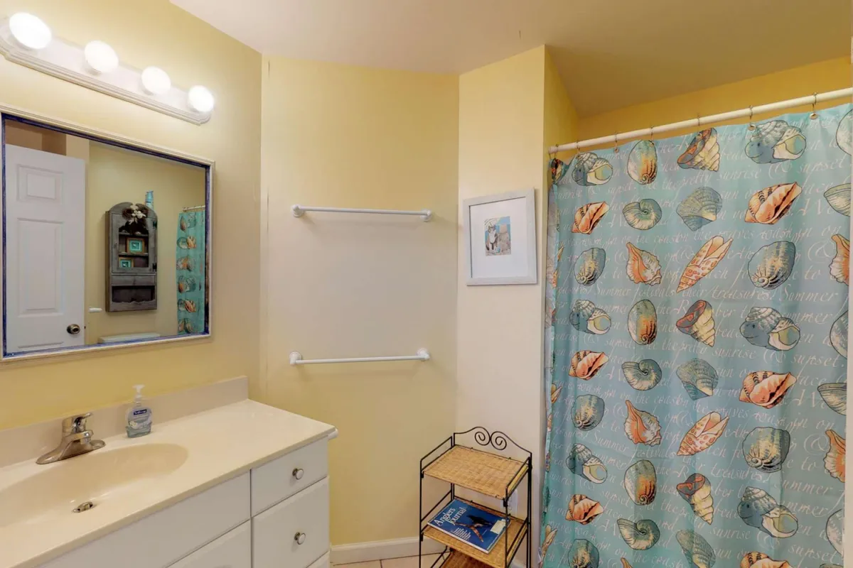 A fresh, inviting bathroom with a vanity sink, mirror, shower curtain, and a rack holding magazines. The space is bright and welcoming, ideal for a refreshing shower or a relaxing soak after a day at the beach.