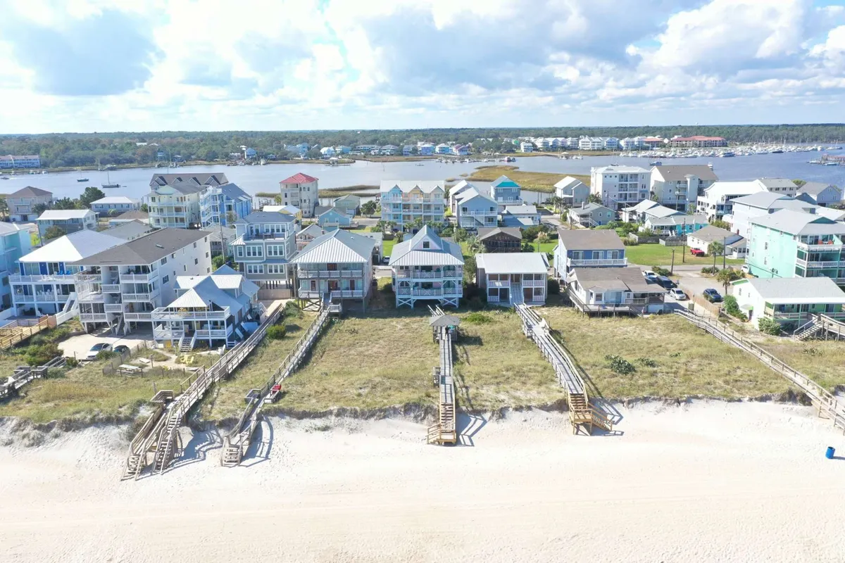 This image showcases a cluster of beachfront houses with wooden walkways leading out over sand dunes, opening to a broad waterfront view. The setting sun casts a warm glow, highlighting opportunities for outdoor gatherings and leisure activities just steps from the property.