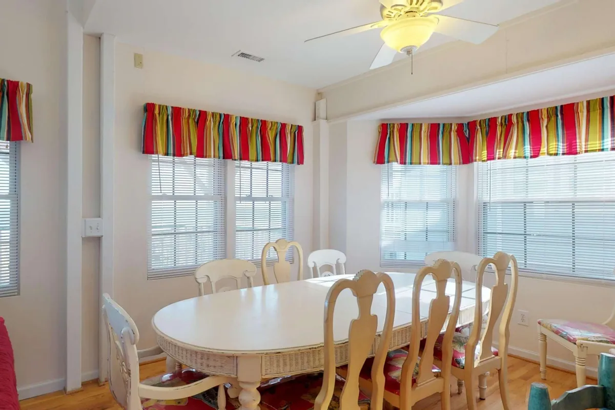 A warm and inviting dining space featuring a large white dining table, colorful window treatments, a ceiling fan, and plenty of space for group meals or board games. Natural light fills the area, creating a cheerful atmosphere.