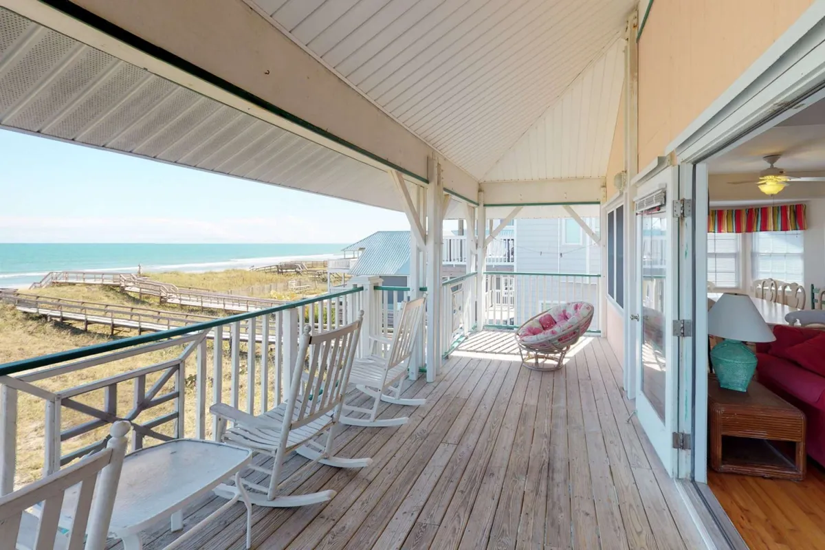 This exterior scene shows a porch with inviting rocking chairs, decorative pillows, and a sweeping view of the beach. The tranquil setting suggests a perfect spot for morning or evening relaxation, reading, or enjoying a drink.