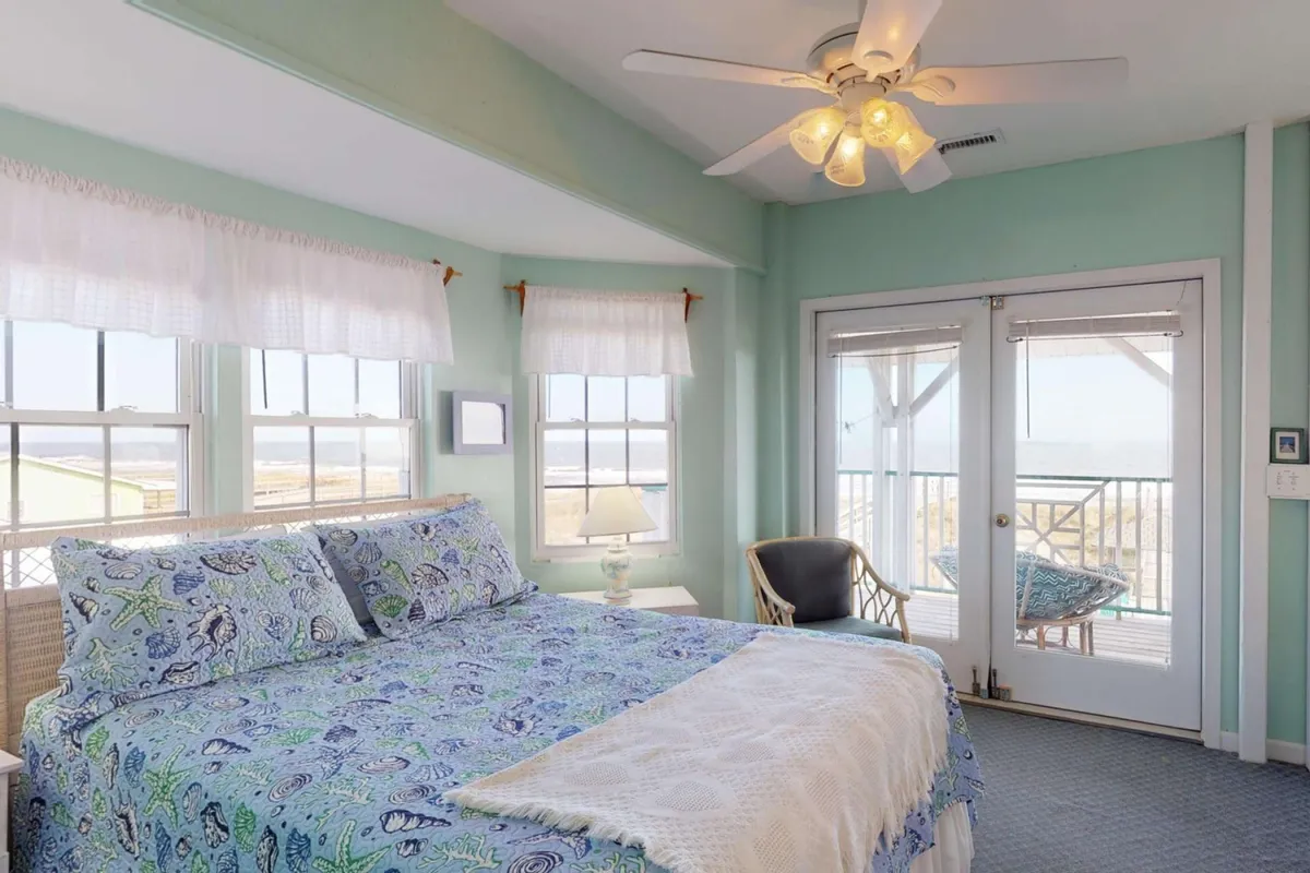 A spacious bedroom featuring a bed with patterned bedding, large windows that let in abundant natural light, and a door leading to a private balcony. The room is designed for comfort and relaxation, with an armchair positioned for reading or enjoying the view.
