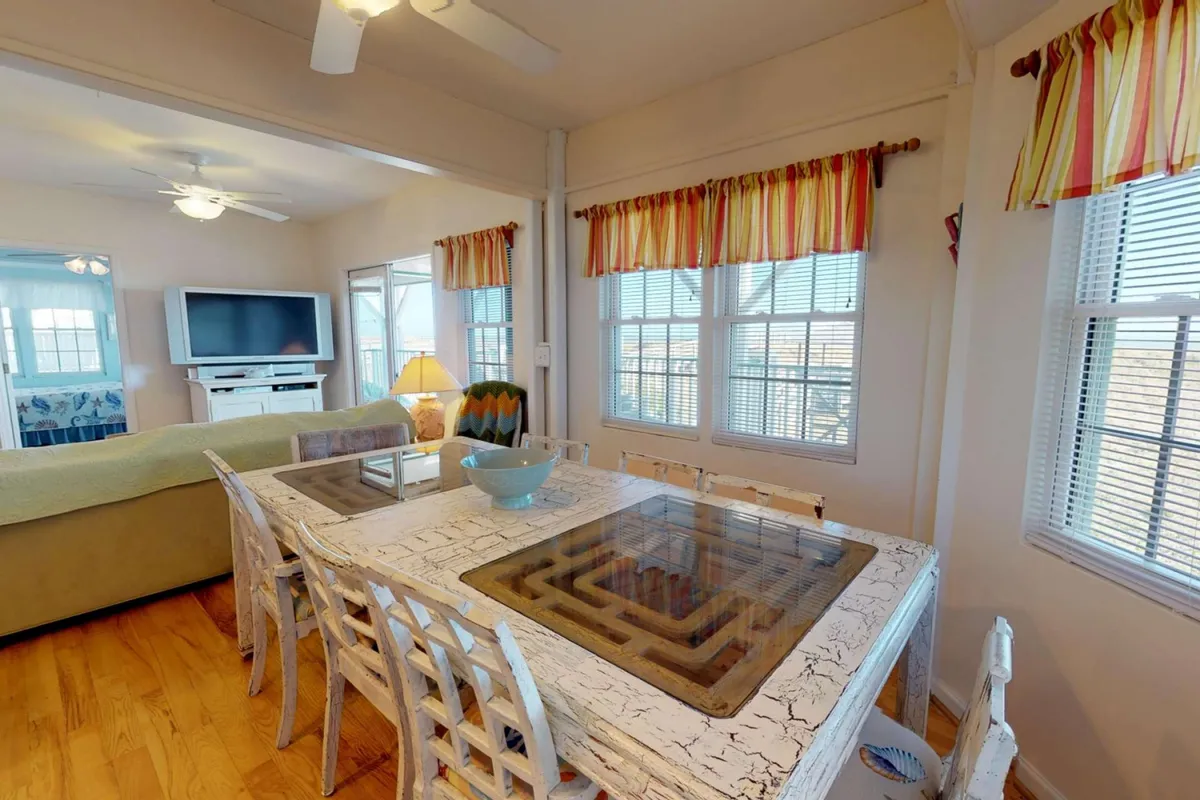 A bright interior scene showcasing a dining table set near large windows, with a casual living area and TV in the background. The space is ideal for family meals, game nights, or relaxed group gatherings after a day of exploring.