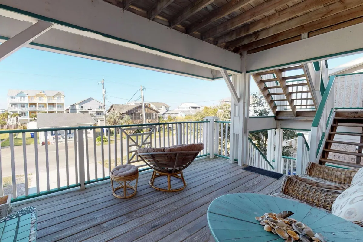 A wooden deck furnished with rattan chairs and a round table offers plentiful seating for groups, with a staircase leading up and an open view—suggesting breezy afternoons, social gatherings, and relaxation. The scene is tranquil and inviting, perfect for group meals or cocktails with a view.